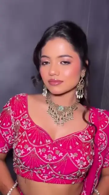 A woman in a hot-pink embroidered blouse with layered silver and stone jewellery, standing against a grey wall.