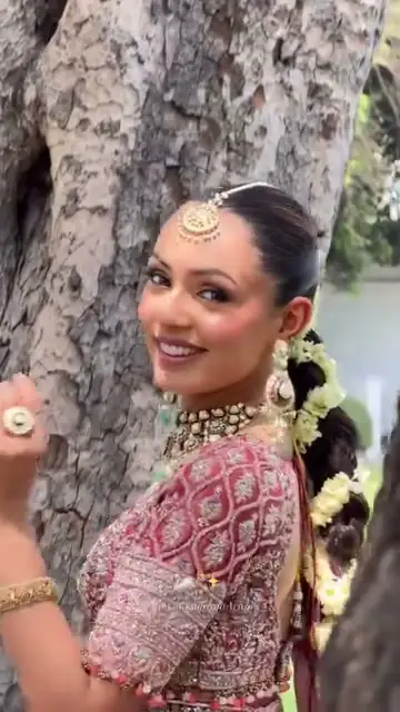 A woman in a beige and gold heavily embellished draped outfit with a diamond choker and jhumka earrings, one hand on her hip on an indoor staircase.